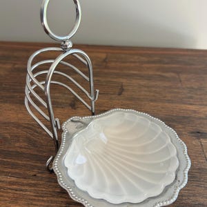 May include: A silver-coloured metal toast rack with a circular handle and a seashell-shaped dish. The dish is white with a silver beaded edge. The rack has a curved design.