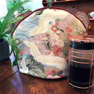 May include: A decorative tea cozy with a crane design, featuring white birds, pink flowers, and blue water elements. A copper and glass French press coffee maker sits next to the cozy on a wooden table.
