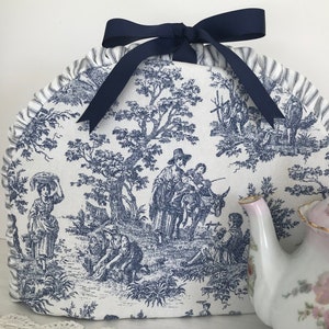 May include: A blue and white toile tea cozy with a navy blue ribbon. The cozy features a pastoral scene and a striped border. A pink and gold floral teapot is partially visible on the right.