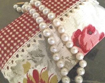 May include: A rectangular fabric pouch featuring a red and white star pattern on one side and a cream-coloured floral print on the other, separated by a white lace trim. A single strand of white pearls is draped across the surface of the pouch.