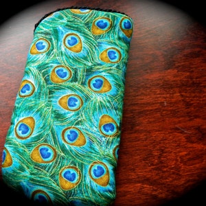 Puede incluir: Una funda rectangular de tela con un patrón repetitivo de plumas de pavo real en tonos turquesa, verde y dorado. La funda tiene una estructura suave y acolchada con un borde negro en la parte superior para guardar objetos pequeños.