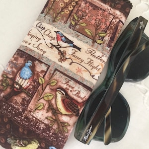 Peut inclure: Un étui à lunettes en tissu brun et beige avec un motif d'oiseaux et de fleurs. L'étui porte l'inscription "Chirp Chirp Singing Song" et "Fluttery Fancy Flight". Une paire de lunettes de soleil noires et vertes en écaille de tortue est posée sur l'étui.