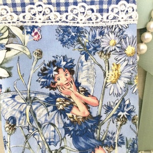Peut inclure: Un tissu bleu et blanc avec un motif floral représentant une fée aux ailes bleues et une couronne de fleurs. La fée se tient les mains au visage et porte une robe bleu clair avec un corsage brun. Le tissu est orné d'une bordure en dentelle blanche en haut.