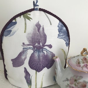May include: A white tea cozy with a purple iris floral print and a brown rope trim. The cozy is sitting on a table next to a white teapot with a pink floral design.