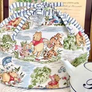 May include: A whimsical tea cozy featuring a charming Winnie the Pooh and friends design. The cozy has a scalloped edge and is adorned with a blue and white striped bow. A white teapot with a blue design is in the foreground.