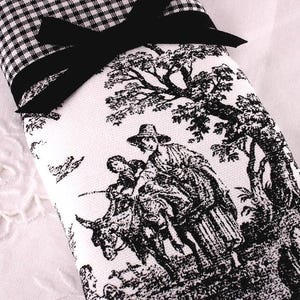 Peut inclure: Une serviette en tissu de style toile de Jouy noir et blanc avec un nœud noir. La serviette présente une scène pastorale avec des personnages montant un âne. Le bord supérieur a un motif vichy noir et blanc.