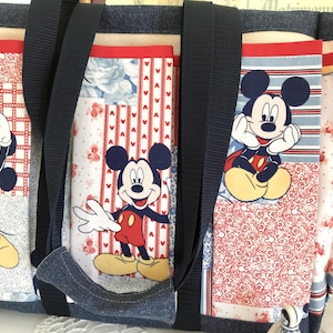 May include: A large tote bag featuring Mickey Mouse designs. The bag has a denim base and black straps. The panels showcase Mickey in various poses against red, white, and blue patterned backgrounds. The bag is designed for carrying items.