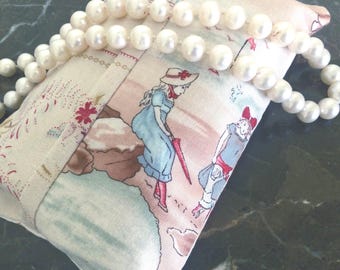 May include: A rectangular fabric sachet featuring a printed illustration of two figures in vintage-style clothing walking on a beach, accented by a strand of white pearls draped across the top surface of the cream-coloured textile.