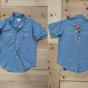 Peut inclure: Une chemise en denim bleu clair à manches courtes et col blanc. La chemise est ornée d'une broderie florale au dos et d'une bordure cloutée argentée autour du col, des poignets et des poches. La chemise a également un "JP" brodé en rose et blanc sur la poitrine gauche.