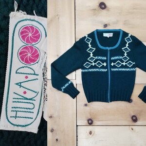 May include: A dark green knitted cardigan with a white and blue geometric pattern. The cardigan has a zipper closure and a label that reads "TRAP" with a pink and green design.