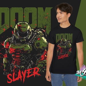 May include: Black t-shirt featuring a green and red graphic of a futuristic armored figure with the word "DOOM" above and "SLAYER" below. The design is distressed, with a grunge aesthetic. The person is wearing blue jeans.