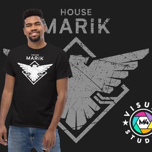 May include: Black t-shirt with a white graphic of a bird inside a diamond shape, and the text "HOUSE MARIK". The shirt is worn by a person in blue jeans. The background features a large, distressed gray eagle graphic and the text "HOUSE MARIK".