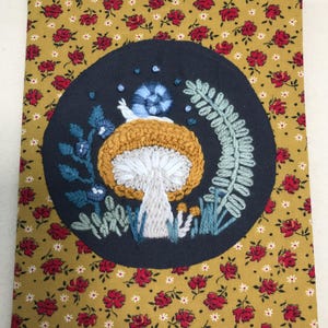 May include: Embroidered journal cover featuring a whimsical mushroom design with a snail, surrounded by foliage. The design is set against a navy blue circle, with a floral pattern background in gold and red. The journal cover is rectangular.