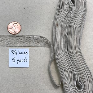 May include: A roll of light gray ribbon, 5/8" wide and 8 yards long, with a delicate floral lace edge. A penny is next to the ribbon for size comparison. The ribbon is neatly coiled on a neutral fabric surface.