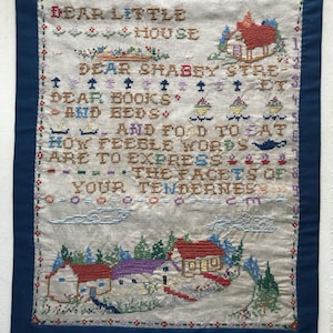 May include: A vintage embroidered sampler with a blue border. The sampler features text and images of houses, trees, and other decorative elements. The text reads "DEAR LITTLE HOUSE, DEAR SHABBY STREET, DEAR BOOKS AND BEDS, AND FOOD TO EAT".