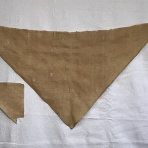 May include: A tan-colored triangular bandana with a matching small square patch. The bandana is made of a soft, textured fabric. The items are displayed on a white surface.