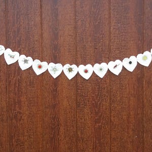 May include: A white heart-shaped garland with various embroidered designs and embellishments. The garland is strung across a dark brown wooden background. Each heart features a unique design, including floral patterns and colorful accents.