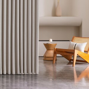 May include: A neutral-toned, vertically-pleated curtain panel dominates the left side of the image. A wooden chair with a woven back and a small side table are visible. A decorative vase and a pillow with a black stripe add to the room's decor.
