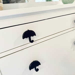 Puede incluir: Cómoda blanca con tres cajones. Cada cajón tiene una manija negra en forma de paraguas.