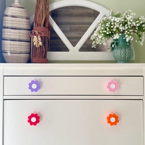 May include: A white dresser with four flower-shaped drawer pulls in purple, pink, red, and orange. The dresser is set against a backdrop of decorative items, including a vase, dried plants, and a green vase with white flowers.