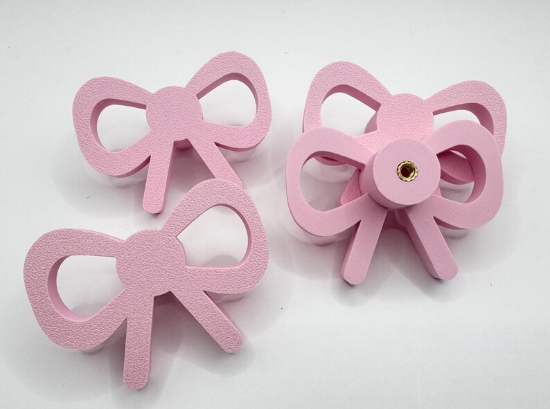 Może przedstawiać: Trzy r&oacute;żowe gałki do szafek w kształcie kokardki. Dwie to pojedyncze kokardy, a jedna ma centralny punkt mocowania. Gałki wykonane są z teksturowanego materiału i mają jasnor&oacute;żowy kolor.