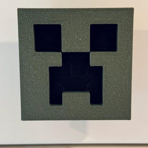May include: A green 3D printed Minecraft Creeper face. The Creeper face is a popular video game character.
