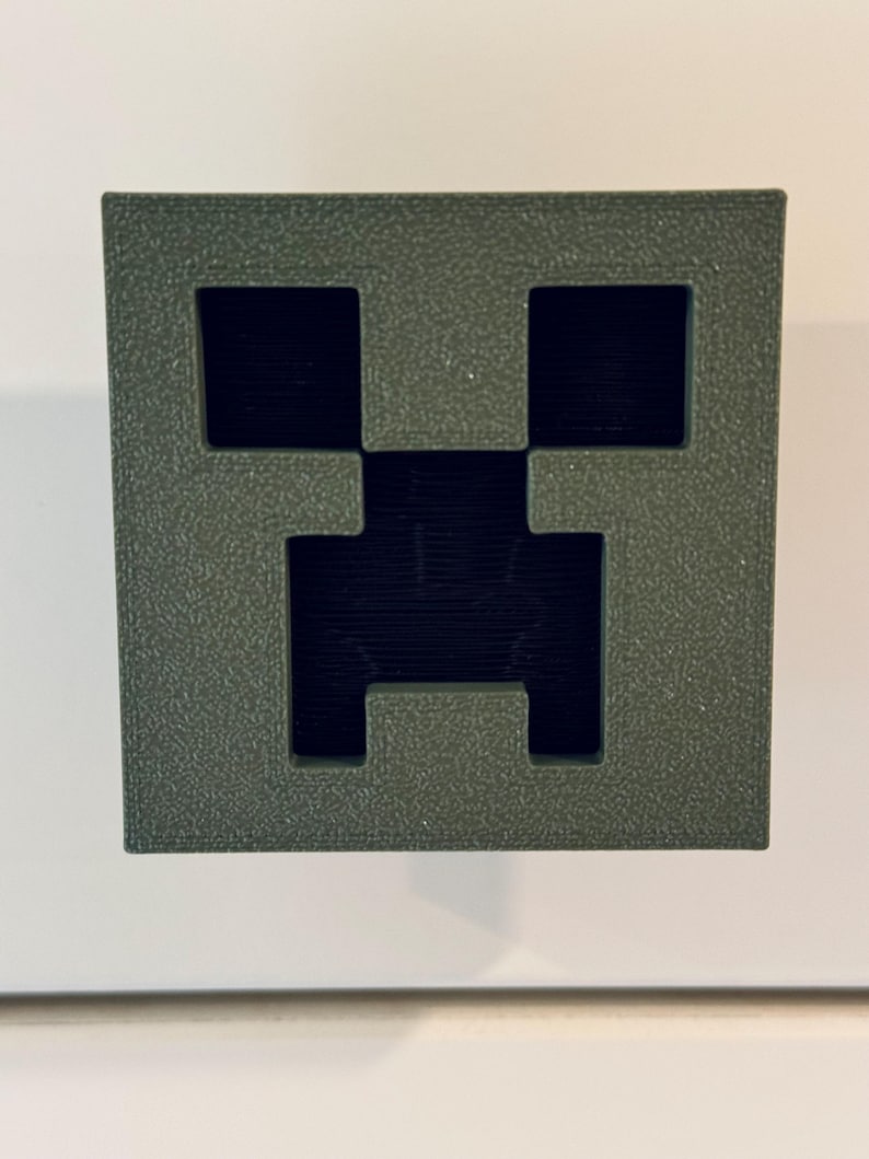 May include: A green 3D printed Minecraft Creeper face. The Creeper face is a popular video game character.