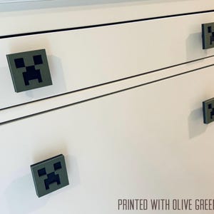 May include: White dresser with four drawers featuring olive green Minecraft Creeper face drawer pulls. "PRINTED WITH OLIVE GREEN COLOR" is printed on the bottom of the dresser.