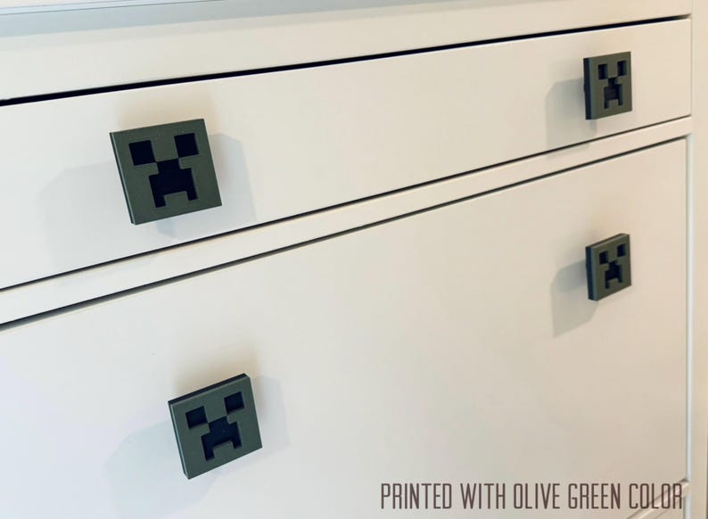May include: White dresser with four drawers featuring olive green Minecraft Creeper face drawer pulls. "PRINTED WITH OLIVE GREEN COLOR" is printed on the bottom of the dresser.