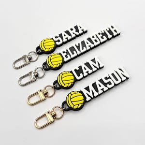 May include: Four black and white personalized keychains with a yellow volleyball graphic. The keychains feature the names "SARA", "ELIZABETH", "CAM", and "MASON".