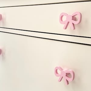 Może przedstawiać: Zbliżenie białej komody z czterema r&oacute;żowymi uchwytami w kształcie kokardki. Uchwyty są r&oacute;wnomiernie rozmieszczone i dodają meblom dekoracyjnego charakteru. Kokardy są w kolorze jasnor&oacute;żowym.