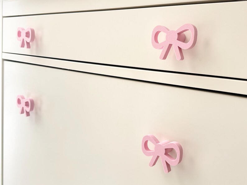 Może przedstawiać: Zbliżenie białej komody z czterema r&oacute;żowymi uchwytami w kształcie kokardki. Uchwyty są r&oacute;wnomiernie rozmieszczone i dodają meblom dekoracyjnego charakteru. Kokardy są w kolorze jasnor&oacute;żowym.