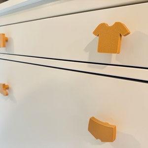 May include: White dresser with four drawers. Each drawer has a different shaped, tan-colored, wooden pull. The pulls are shaped like a t-shirt, a pair of pants, a pair of underwear, and a shirt.