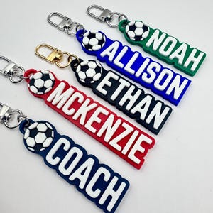 May include: A collection of personalized soccer keychains in various colors, including green, blue, red, and black. Each keychain features a soccer ball design and a name or word in white lettering. The keychains are attached to silver or gold-colored clasps.