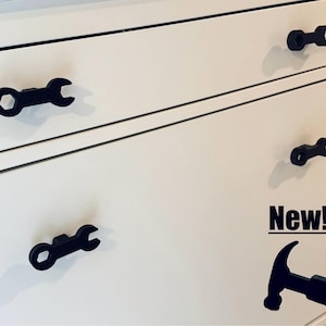May include: Four black wrench-shaped drawer pulls are mounted on a white dresser. The top drawer has the text "New!" written on it.
