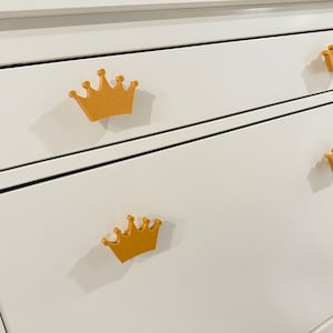 May include: White dresser with three drawers featuring gold crown-shaped drawer pulls.