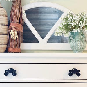 May include: White dresser with two drawers. The top drawer has a black monster truck pull handle. The bottom drawer has a black monster truck pull handle. The dresser is decorated with a vase of white flowers, a brown wicker basket, and a brown and white ceramic vase.