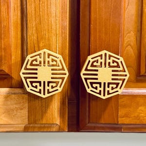 May include: Two gold-colored, octagonal cabinet knobs with an intricate geometric design. The knobs are mounted on a pair of wooden cabinet doors with a warm, reddish-brown finish. The knobs are centered on each door.
