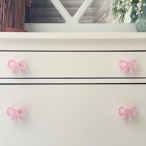 May include: A white dresser featuring four light pink bow-shaped drawer knobs. Each knob is shaped like a tied ribbon with two loops and two hanging ends, mounted centrally on the front of each drawer to serve as a functional handle.
