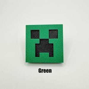 May include: A green square with a black Creeper face from the video game Minecraft. The Creeper face is a pixelated design with a square mouth and eyes.