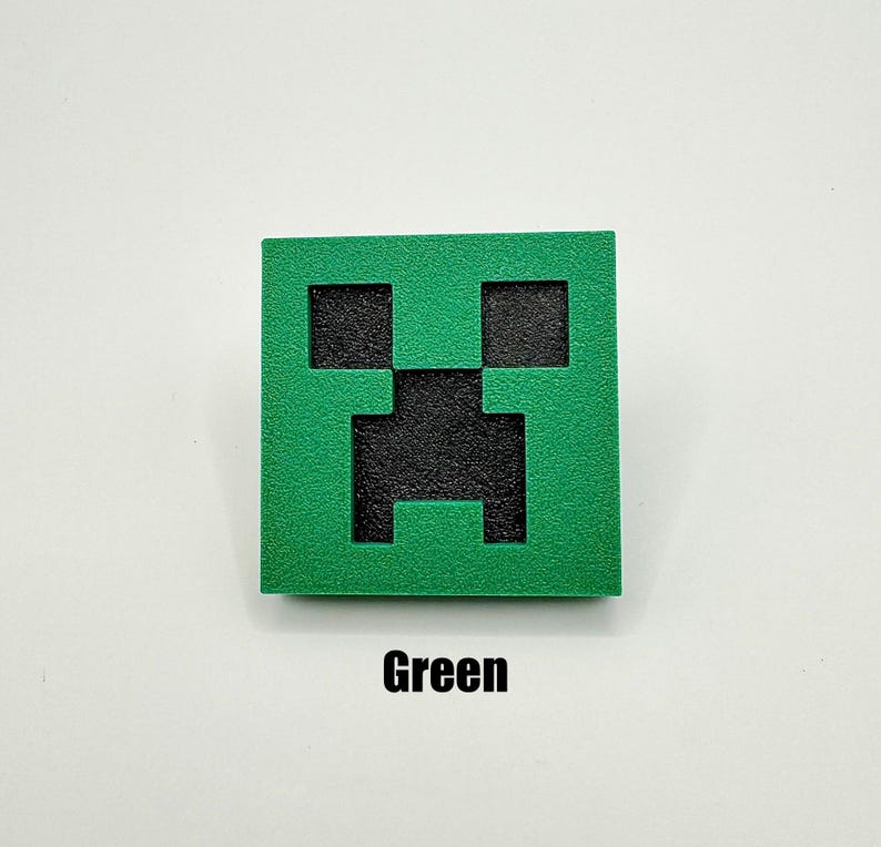 May include: A green square with a black Creeper face from the video game Minecraft. The Creeper face is a pixelated design with a square mouth and eyes.