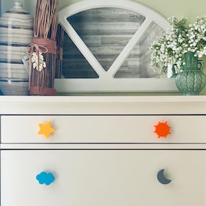 May include: A white dresser with four colorful drawer knobs shaped like a yellow star, orange sun, blue cloud, and gray crescent moon. The dresser is decorated with a vase of flowers and other decorative items.