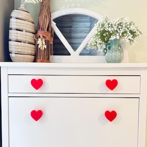 May include: White dresser with four red heart-shaped drawer pulls. The dresser has two drawers and a cabinet door. The dresser is decorated with dried flowers and a vase of white flowers.