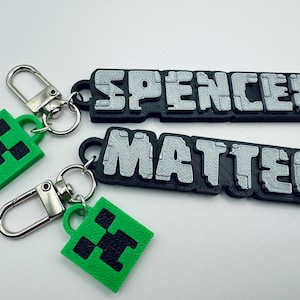 May include: Two black and silver keychains with the names "Spencer" and "Matteo" printed on them. Each keychain has a green charm with a black Minecraft Creeper face.