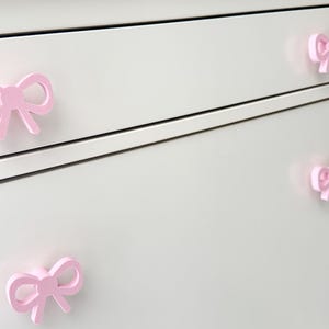 Może przedstawiać: Zbliżenie białej komody z czterema r&oacute;żowymi uchwytami w kształcie kokardek. Uchwyty są r&oacute;wnomiernie rozmieszczone i dodają meblom dekoracyjnego charakteru. Kokardy mają delikatny r&oacute;żowy kolor.