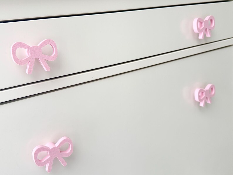 Może przedstawiać: Zbliżenie białej komody z czterema r&oacute;żowymi uchwytami w kształcie kokardek. Uchwyty są r&oacute;wnomiernie rozmieszczone i dodają meblom dekoracyjnego charakteru. Kokardy mają delikatny r&oacute;żowy kolor.