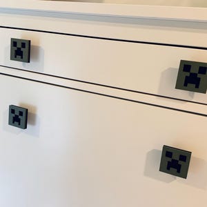 May include: White dresser with four drawers. Each drawer has a black and green Minecraft Creeper face pull.