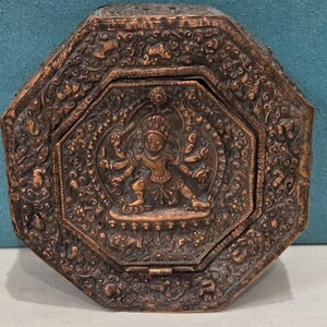 May include: An octagonal copper-toned metal box featuring an intricate embossed relief of a multi-armed deity at the center. The surface is covered in detailed scrollwork and floral patterns, with a hinged lid and a dark, weathered finish.
