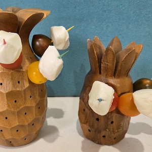 May include: Two wooden pineapple-shaped serving dishes with small white cheese balls and red and yellow cherry tomatoes on skewers.