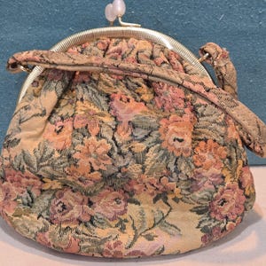 May include: A vintage floral tapestry coin purse with a gold-tone metal frame and clasp. The purse features a repeating pattern of pink, peach, and green flowers on a cream background. It has a small handle and a decorative clasp with a pearl-like accent.